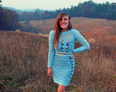 a woman standing in a field wearing a blue crochet dress and smiling at the camera