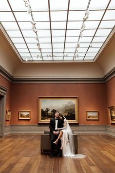 a bride and groom sitting on a bench in the middle of a room with paintings