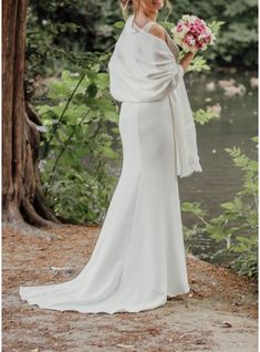 a woman in a white dress holding a bouquet and standing next to a river wearing a shawl