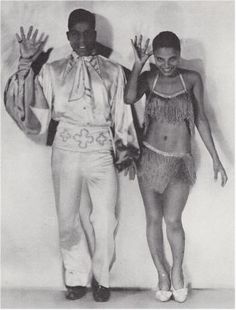 an old black and white photo of two people dressed in native american dance costumes, one holding his hands up