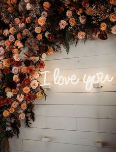 i love you neon sign surrounded by flowers and greenery on a white wooden wall