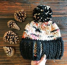 a hand is holding a knitted hat with pine cones on the table next to it