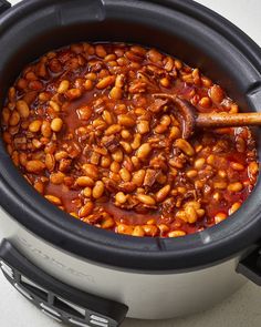 a crock pot filled with baked beans on top of a white counter next to a wooden spoon