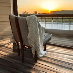 a chair with a blanket on it sitting on a porch next to a lake at sunset