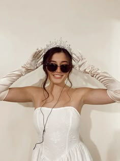 a woman wearing sunglasses and a tiara with her hands on her head, standing in front of a white wall