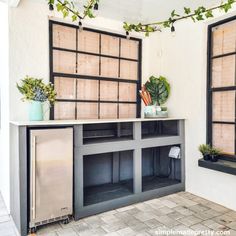 an outdoor kitchen with plants on the counter and two windows above it that have bamboo shades over them