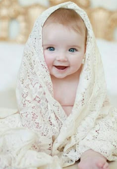 a baby wearing a white shawl on top of a bed smiling at the camera