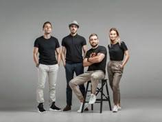 four people standing and sitting on stools in front of a gray background with one person holding a cell phone