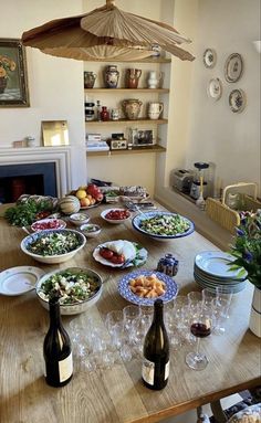 a wooden table topped with lots of plates and bowls filled with food next to bottles of wine