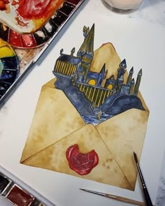 an envelope with a castle drawn on it next to paintbrushes and watercolors