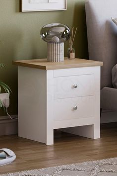 a white night stand with two drawers in front of a couch and potted plant