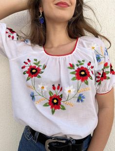 a woman wearing a white top with red and blue flowers on it, leaning against a wall