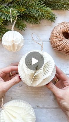 two hands are holding paper balls and twine on a white wooden table with pine branches