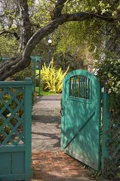 a green gate is open on a brick walkway in front of a tree and shrubbery