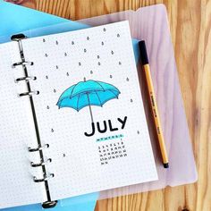 a notebook with the word july written on it next to a pen and an umbrella