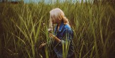 a woman standing in tall grass with her face close to the ground and looking at flowers