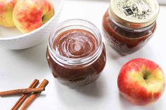 apples and cinnamon sticks sit next to two jars of apple jam on a white table