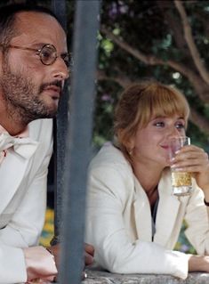 a man and woman sitting at a table with drinks in front of them, both looking off into the distance