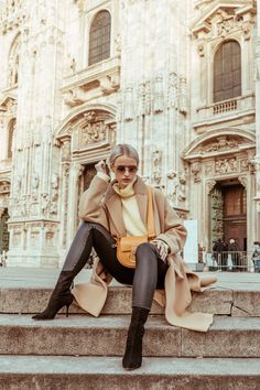 a woman sitting on the steps in front of a building wearing black boots and a tan coat