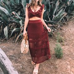 a woman standing in front of some plants holding a handbag and looking at the camera