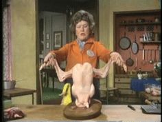 an old woman standing in front of a fake chicken on a table with other items around her