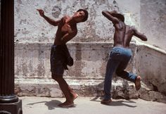 two men are dancing in front of a stone wall and one has his arms stretched out