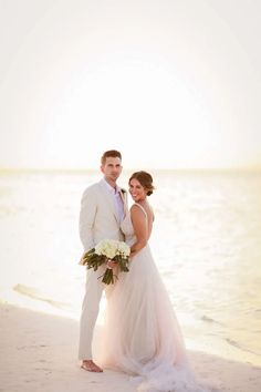 a bride and groom standing on the beach at sunset in front of the ocean with their bouquet