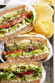 three sandwiches with lettuce, tomatoes and other toppings next to potato chips