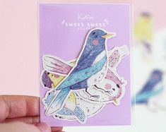 a hand holding up a purple card with a bird on the front and two cats on the back