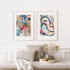 two paintings hang on the wall above a bench in a white room with pillows and vases