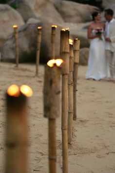 a couple standing next to each other on top of a sandy beach with candles in the sand