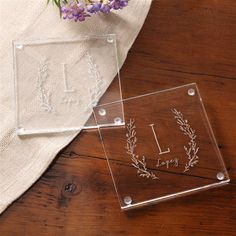 two clear acrylic coasters sitting on top of a wooden table next to purple flowers