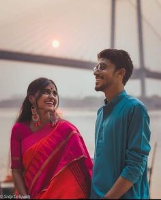 a man and woman standing next to each other in front of a bridge at sunset