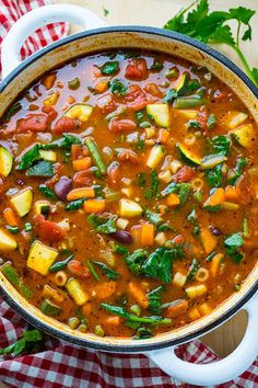 a large pot filled with vegetable soup on top of a checkered cloth next to a spoon