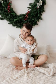 two children are sitting on a bed with christmas wreaths behind them and one is holding the baby