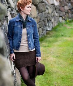 a woman with red hair wearing a denim jacket and black tights, leaning against a stone wall