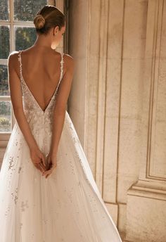 a woman in a white wedding dress standing by a window with her back to the camera