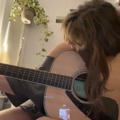 a woman sitting on the floor playing an acoustic guitar in front of a window with a plant behind her