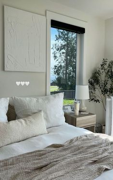 a bed with white pillows and blankets in front of a window that has a painting on the wall