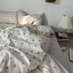 an unmade bed with white sheets and pink flowers on it, next to a lamp