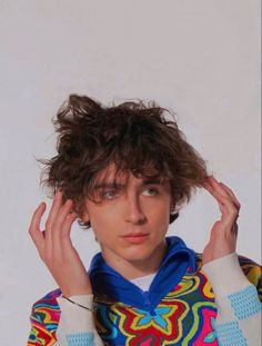 a young man with curly hair wearing a colorful sweater and holding his hands up to his head