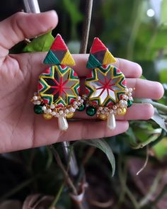 a pair of colorful earrings is held in someone's hand with greenery behind it