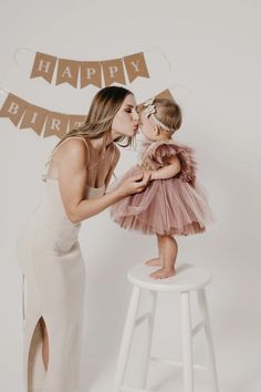 a mother kissing her daughter's cheek while standing on a stool in front of a happy birthday banner