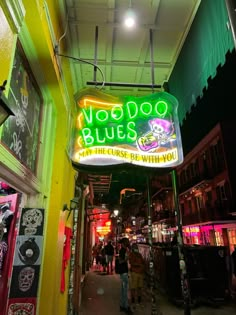 the neon sign for voodoo blues is lit up in green and yellow colors, with people walking down the street below