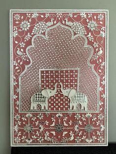 an intricately designed card with elephants and birds on red paper, in front of a gray wall