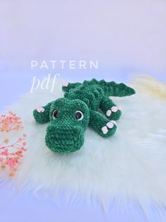 a green crocheted alligator laying on top of a white fur rug next to a pink flower