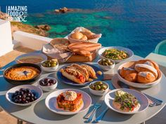 a table full of food on top of a patio next to the ocean with blue water in the background