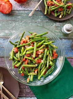 green beans and tomatoes in a glass bowl on a wooden table with silverware next to it