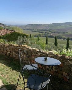 an outdoor table and chairs on the side of a stone wall with rolling hills in the background