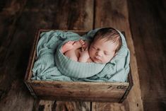 a newborn baby is curled up in a wooden box on a wood floor with his head tucked under the blanket